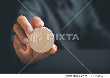 A hand holds a blank round wooden sign in front of a blurred background. . A hand holds a blank round wooden sign in front of a blurred background. . 134983206