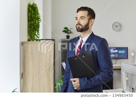 Confident businessman in suit with clipboard standing in modern corporate office Confident businessman in suit with clipboard standing in modern corporate office 134983450
