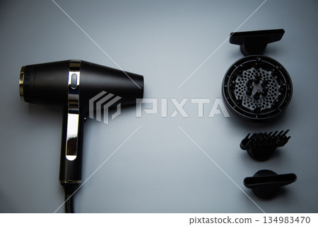 A black hair dryer on one side, on the other there are different drying attachments - a comb, a diffuser and classic nozzles of different diameters on a gray background, top view. The concept of A black hair dryer on one side, on the other there are different drying attachments - a comb, a diffuser and classic nozzles of different diameters on a gray background, top view. The concept of 134983470