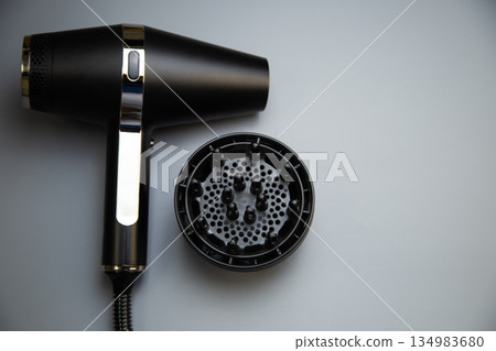 On one side there is a black hair dryer and a diffuser nozzle on a gray background, a top view with space for text. The concept of hairdressing, hair styling, self-care. High quality photo On one side there is a black hair dryer and a diffuser nozzle on a gray background, a top view with space for text. The concept of hairdressing, hair styling, self-care. High quality photo 134983680
