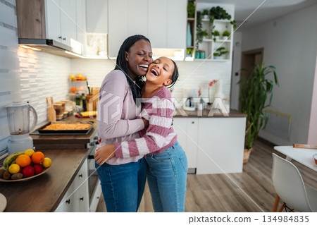 Mother and daughter embracing and laughing in kitchen 134984835