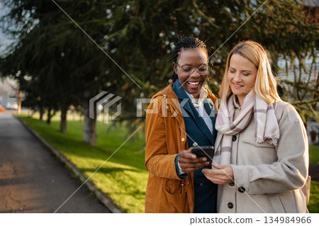 Diverse women friends smiling using smartphone outdoors Diverse women friends smiling using smartphone outdoors 134984966