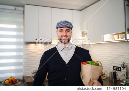Smiling man holding groceries bag in modern kitchen Smiling man holding groceries bag in modern kitchen 134985104