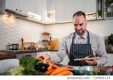 Man in apron preparing fresh vegetables following recipe book Man in apron preparing fresh vegetables following recipe book 134985114
