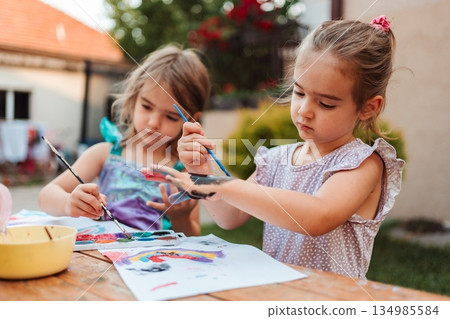 Two preschool girls painting with fingers and brushes in backyard Two preschool girls painting with fingers and brushes in backyard 134985584