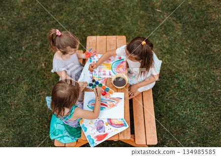 Three little girls painting rainbows at a wooden table in the garden 134985585