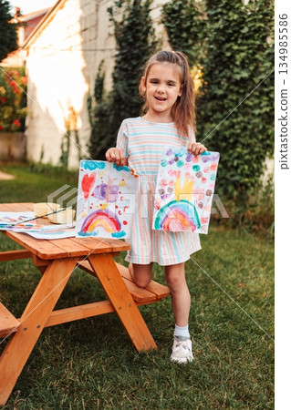 Little Girl Showing Her Colorful Drawings in the Garden Little Girl Showing Her Colorful Drawings in the Garden 134985586