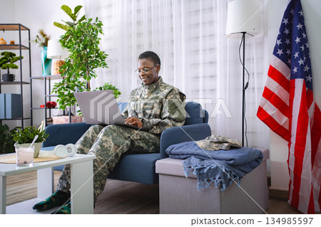 Female soldier sitting on sofa using laptop with United States flag in background Female soldier sitting on sofa using laptop with United States flag in background 134985597