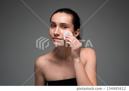 Young woman using cotton pad on face in studio, clean skincare routine concept, natural beauty portrait for cosmetic and facial care advertising 134985612