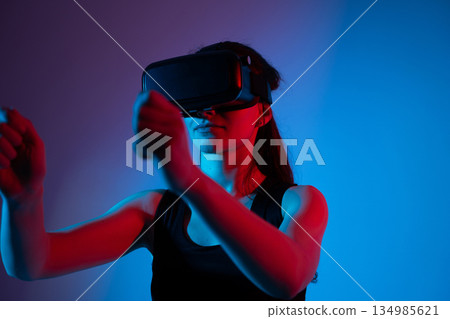 Young woman wearing VR headset making forward hand gesture in immersive virtual environment Young woman wearing VR headset making forward hand gesture in immersive virtual environment 134985621