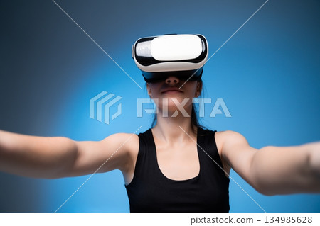 Young woman wearing VR headset with arms extended forward, immersive virtual reality interaction on blue studio background 134985628