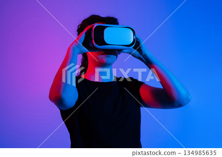Woman adjusting virtual reality headset with both hands while preparing for immersive digital experience in neon studio lighting Woman adjusting virtual reality headset with both hands while preparing for immersive digital experience in neon studio lighting 134985635