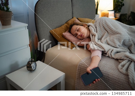 Woman sleeping in bed holding smart phone in bedroom Woman sleeping in bed holding smart phone in bedroom 134985711