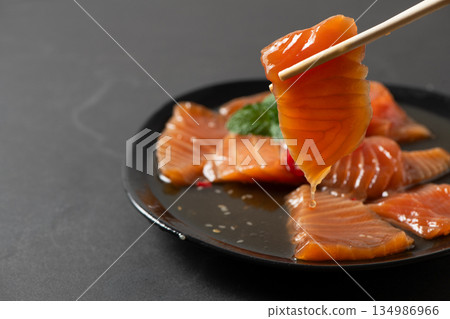 orean soy marinated salmon slices being picked with chopsticks, sauce dripping on black plate. Popular Korean food known as salmon jang, premium seafood orean soy marinated salmon slices being picked with chopsticks, sauce dripping on black plate. Popular Korean food known as salmon jang, premium seafood 134986966