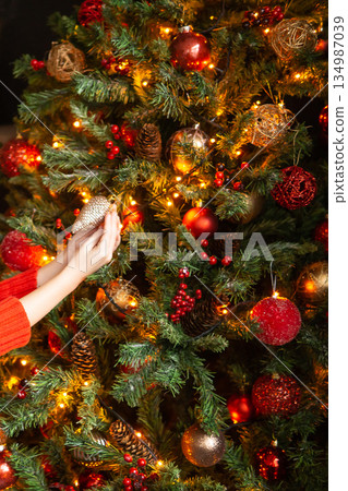 Child decorating Christmas tree with ornaments. Kid hand hanging red festive bauble on fir branch. Holiday season celebration concept. 134987039