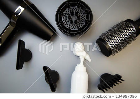 Top view of hair styling foam in a white bottle lies on a gray background surrounded by hair drying attachments, a hairdryer and a round styling comb. The concept of long-term hair styling, creating Top view of hair styling foam in a white bottle lies on a gray background surrounded by hair drying attachments, a hairdryer and a round styling comb. The concept of long-term hair styling, creating 134987080