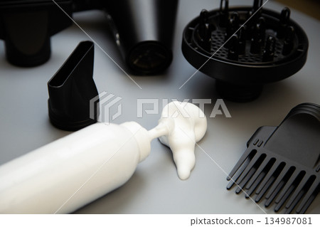 Hair styling foam in a white bottle lies on a gray background, surrounded by hair drying nozzles and a hairdryer. The concept of long-term hair styling, creating hairstyles, curls and taking care of Hair styling foam in a white bottle lies on a gray background, surrounded by hair drying nozzles and a hairdryer. The concept of long-term hair styling, creating hairstyles, curls and taking care of 134987081