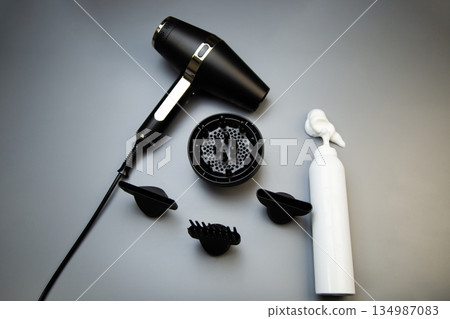 A black hair dryer with different attachments - a diffuser for creating curls and curls, a comb and classic lie on a gray background next to a hair mousse for long-lasting styling with white foam on A black hair dryer with different attachments - a diffuser for creating curls and curls, a comb and classic lie on a gray background next to a hair mousse for long-lasting styling with white foam on 134987083