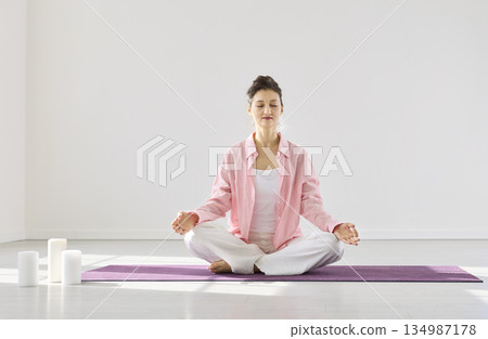Peaceful woman meditating in lotus on purple mat with candles in bright studio 134987178