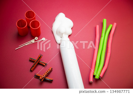 Close-up view from above in the center of a bottle of foam for long-lasting hair styling lies on a pink background, white foam has poured out of it, different types of curlers lie around. The concept Close-up view from above in the center of a bottle of foam for long-lasting hair styling lies on a pink background, white foam has poured out of it, different types of curlers lie around. The concept 134987771