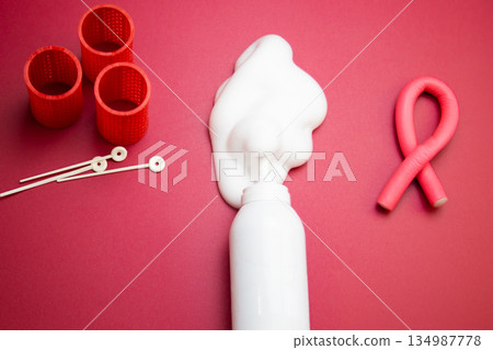 Close-up view from above in the center of a bottle of foam for long-lasting hair styling lies on a pink background, white foam poured out of it, round curlers and a curved pink curler lie nearby. The 134987778