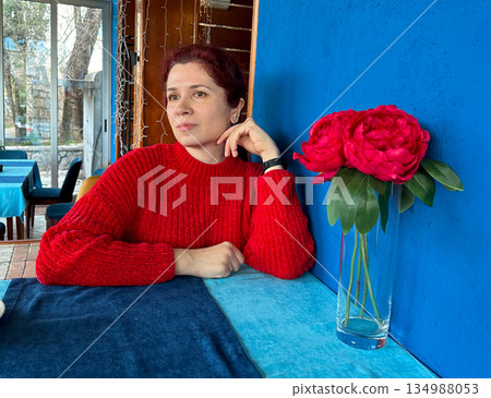 Woman resting her chin at cafe table beside flowers and blue wall. Calm femininity, thoughtful mood, color harmony, and serene lifestyle portrait in cozy interior. 134988053