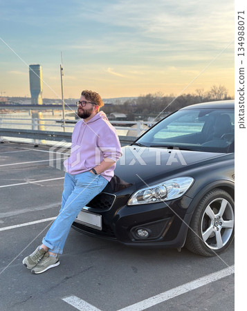 Man leaning on car near river at sunset. Urban lifestyle, freedom, modern masculinity, and reflective pause in city environment Man leaning on car near river at sunset. Urban lifestyle, freedom, modern masculinity, and reflective pause in city environment 134988071