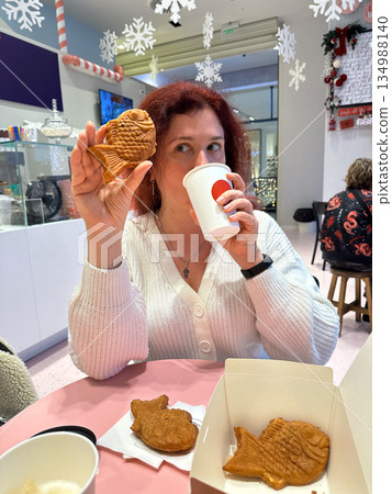 Woman tasting festive drink while holding taiyaki fish shaped pastry. Cozy cafe leisure, playful holiday treat, winter sweetness, relaxed enjoyment, and cheerful christmas atmosphere indoors. 134988140