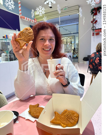 Woman smiling while holding fish shaped taiyaki pastry and warm drink. Playful winter snack, holiday cafe mood, joyful indulgence, cozy atmosphere, and cheerful seasonal lifestyle moment indoors. Woman smiling while holding fish shaped taiyaki pastry and warm drink. Playful winter snack, holiday cafe mood, joyful indulgence, cozy atmosphere, and cheerful seasonal lifestyle moment indoors. 134988155