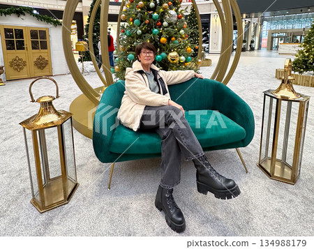 Woman sitting on sofa near decorated Christmas tree installation. Seasonal interior design, festive public space, winter comfort, calm leisure, and lifestyle pause within holiday shopping environment. Woman sitting on sofa near decorated Christmas tree installation. Seasonal interior design, festive public space, winter comfort, calm leisure, and lifestyle pause within holiday shopping environment. 134988179