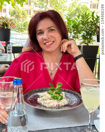 Smiling woman sitting at restaurant table with pasta dish and drinks outdoors. Casual dining portrait, social leisure moment and relaxed lifestyle enjoyment 134988199