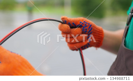 An electrician worker holds a red and black high-voltage electric cable, checks the quality of the cables. Connecting electrical cables. Close up of male hands in protective gloves holding cable. An electrician worker holds a red and black high-voltage electric cable, checks the quality of the cables. Connecting electrical cables. Close up of male hands in protective gloves holding cable. 134988711