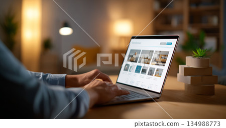 Person browsing furniture website on laptop in cozy living room with warm lighting and modern decor 134988773