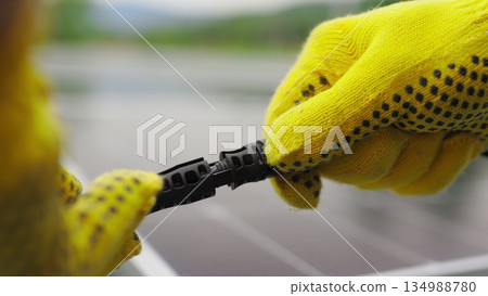 Solar Station Equipment. Connecting wires of solar panels at a solar farm. Engineers connect electrical system outdoors on a background of solar panels. Worker in protective gloves connects wire. Solar Station Equipment. Connecting wires of solar panels at a solar farm. Engineers connect electrical system outdoors on a background of solar panels. Worker in protective gloves connects wire. 134988780