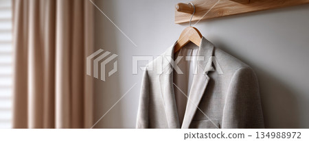 Beige textured blazer hanging on wooden hanger against light wall with soft natural window light 134988972