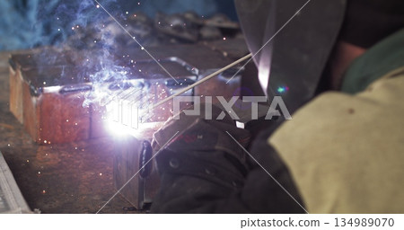 Close-up blacksmith welder in protective mask works with metal using a welding machine, bright sparks and flashes in super slow motion. 134989070
