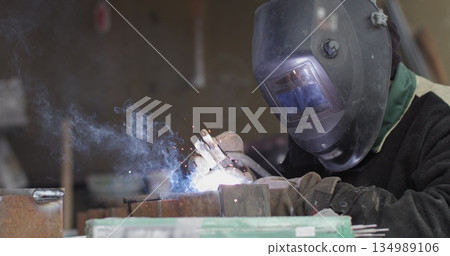 Close-up blacksmith welder in protective mask works with metal steel and iron using a welding machine, bright sparks and flashes in extreme slow motion. 134989106