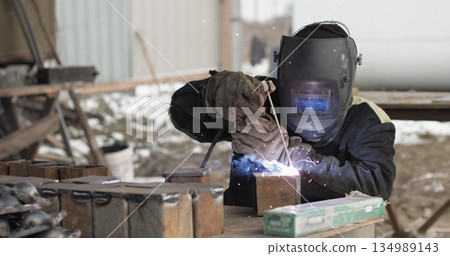 Close-up blacksmith welder in protective mask works with metal using a welding machine, bright sparks 134989143