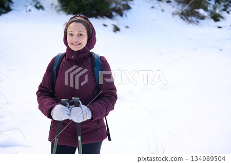 Woman Hiker in Snowy Landscape Wearing Maroon Jacket With Backpack and Trekking Poles 134989504