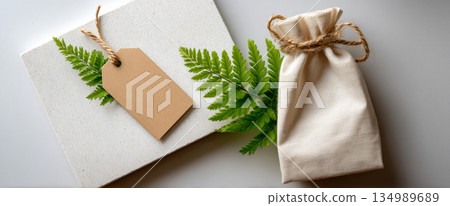 Eco-friendly gift packaging with natural fern leaves, blank kraft paper tag, and beige cotton drawstring bag on textured white surface 134989689