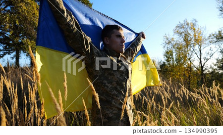 Military man in uniform stands with raised over head flag of Ukraine at countryside. Male ukrainian army soldier with lifted national banner in honor of victory against russian aggression. End of war 134990395