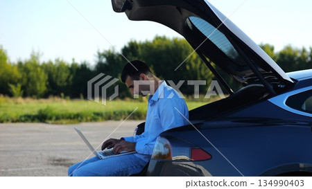 Young handsome entrepreneur works on notebook sitting in electric car trunk. Successful businessman in formal wear using computer at auto. Confident manager typing text on laptop outdoor. Slow mo Young handsome entrepreneur works on notebook sitting in electric car trunk. Successful businessman in formal wear using computer at auto. Confident manager typing text on laptop outdoor. Slow mo 134990403