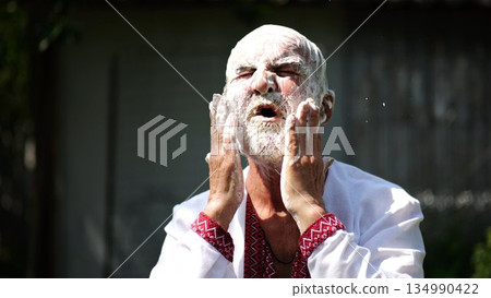 Elderly man wearing in traditional embroidered shirt washing his gray beard fully covered in soap at summer garden. Scene captures care with sense of peace and natural simplicity. Daily ritual concept Elderly man wearing in traditional embroidered shirt washing his gray beard fully covered in soap at summer garden. Scene captures care with sense of peace and natural simplicity. Daily ritual concept 134990422