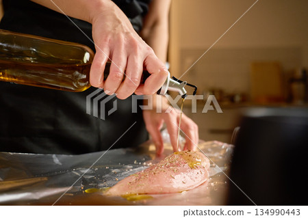 Woman pouring olive oil on raw chicken breast on kitchen table Woman pouring olive oil on raw chicken breast on kitchen table 134990443