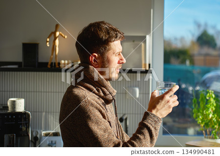 Man drinking water from glass in kitchen interior with daylight Man drinking water from glass in kitchen interior with daylight 134990481