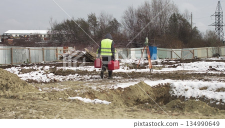 Worker Professional Surveying Engineer At the construction site in his hands two red suitcases. Worker using a theodolite geodetic optical device to measure angles in the horizontal and vertical Worker Professional Surveying Engineer At the construction site in his hands two red suitcases. Worker using a theodolite geodetic optical device to measure angles in the horizontal and vertical 134990649