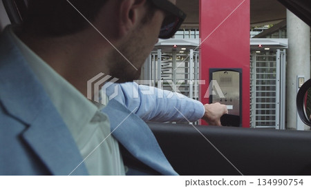 young male businessman in nice suit and glasses paying for parking by bank card with car leaving his office. Office and parking security. young male businessman in nice suit and glasses paying for parking by bank card with car leaving his office. Office and parking security. 134990754