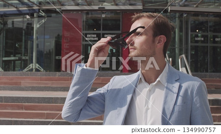 Good-looking confident purposeful adult bearded businessman in glasses in stylish suit going down on steps while leaving the office building and looking at camera. Good-looking confident purposeful adult bearded businessman in glasses in stylish suit going down on steps while leaving the office building and looking at camera. 134990757