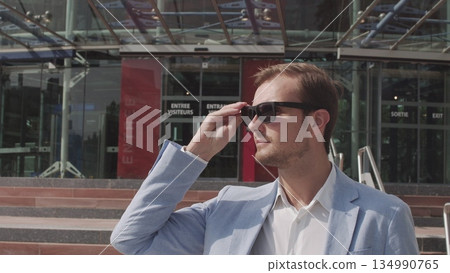 Good-looking confident purposeful adult bearded businessman in glasses in stylish suit going down on steps while leaving the office building and looking at camera. Good-looking confident purposeful adult bearded businessman in glasses in stylish suit going down on steps while leaving the office building and looking at camera. 134990765