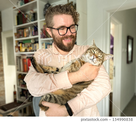 Man holding cat indoors at home environment. Human animal bond, pet companionship, domestic comfort and everyday emotional connection Man holding cat indoors at home environment. Human animal bond, pet companionship, domestic comfort and everyday emotional connection 134990838